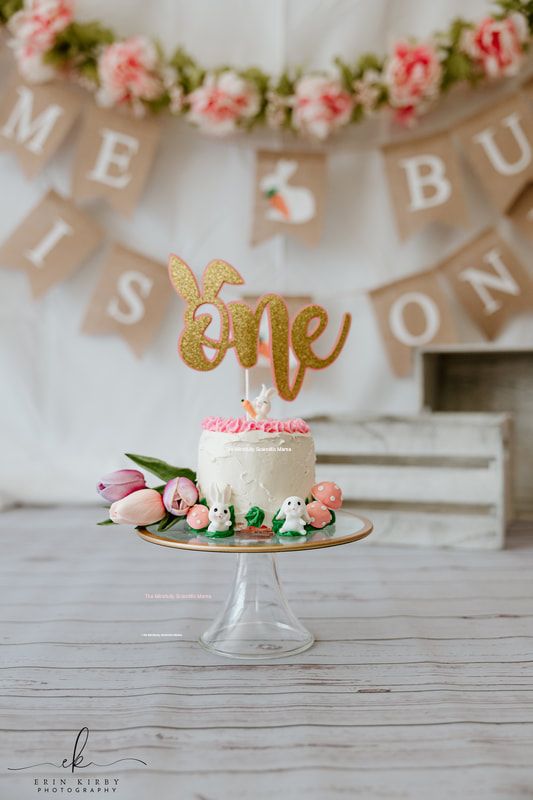 A small white cake in the center of the frame. The cake has a pink frosted border on the top, plus a small white rabbit holding and orange carrot, and a stick holding the word 