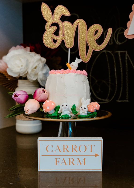 A small white cake in the center of the frame. The cake has a pink frosted border on the top, plus a small white rabbit holding and orange carrot, and a stick holding the word 
